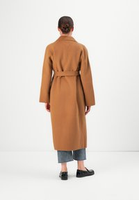 Donna con un cappotto marrone con cintura, jeans blu a vita alta e mocassini neri, in piedi di fronte a un muro bianco semplice, capelli raccolti in un bun.