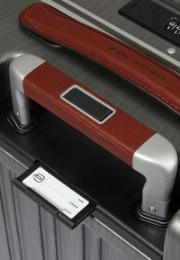 Grijze reiskoffer met gestructureerde afwerking, zilveren metalen accenten, bruine leren handgreep en een afneembare zwarte identificatietag.