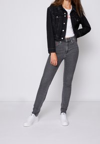 Chaqueta de mezclilla negra sobre una camiseta blanca con texto rojo, combinada con jeans ajustados grises y zapatillas blancas, mostrando un diseño entallado.