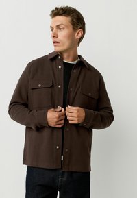 Chaqueta marrón de botones, hecha de un tejido texturizado, con dos bolsillos frontales y un cuello. Llevada sobre una camisa negra.