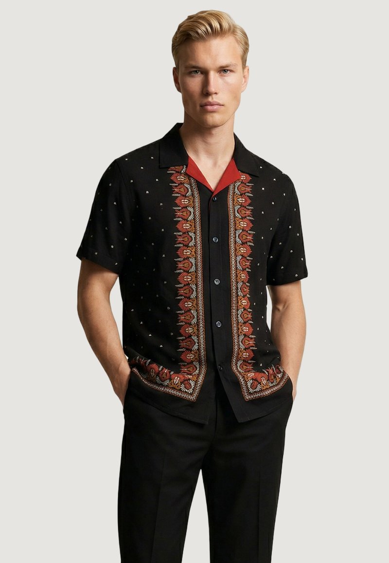 Hombre rubio que lleva una camisa negra de manga corta con rayas verticales florales rojas y naranjas y pequeños puntos blancos, manos en los bolsillos, fondo neutro.