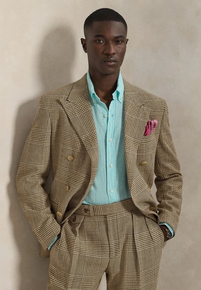 Chaqueta de traje cruzada de cuadros marrones sobre una camisa de lino azul claro, con un pañuelo rosa en el bolsillo y pantalones plisados. Textura suave.