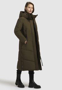 Dlouhá olivově zelená zateplená bunda s vysokým límcem, předním zipem a bočními kapsami na zip. Obsahuje knoflíky podél lemu a strukturovaný povrch.