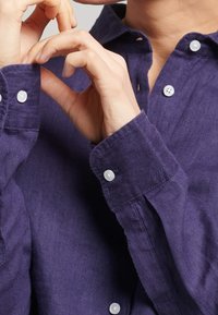 Person som knäpper vita manschetter på en långtärmad, mörk lila skjorta, med fokus på händer och överkropp.