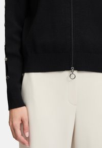 Cardigan en tricot noir avec une fermeture éclair à l'avant, des poignets côtelés et des boutons sur les manches. Associe avec un pantalon beige, montrant une texture lisse.