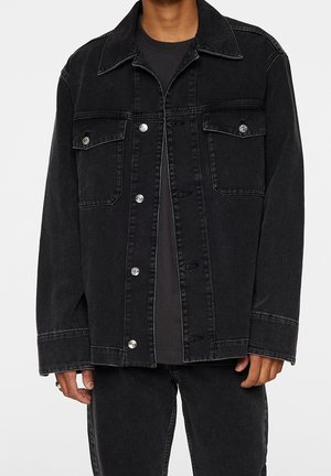 Uomo che indossa una giacca di jeans nera larga con bottoni argentati e due tasche con patta sul petto sopra una camicia nera e pantaloni neri.