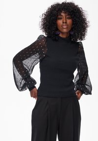 Black top with ribbed fabric and sheer puff sleeves featuring a patterned design. Model wears black trousers, showcasing a fitted silhouette.