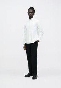 Homme aux cheveux tressés portant une chemise blanche boutonnée et un pantalon noir, debout avec une main dans la poche contre un fond blanc uni.