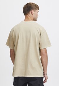 Khaki bomulls t-skjorte med korte ermer og rett snitt. Har rund hals og synlige sømmer langs nedre kant og skuldre.