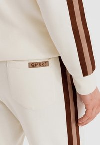Pantalon de survêtement et haut couleur crème avec des rayures marron et beige sur les manches et les côtés du pantalon, avec un patch logo sur la poche arrière.