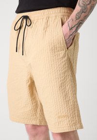 Beige ribbed shorts with an elastic waistband and adjustable black drawstring, featuring side pockets and a small embossed logo.