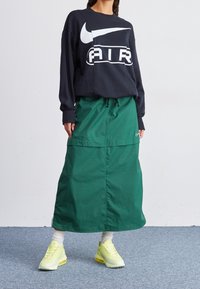 Svart, långärmad tröja med rund halsringning och vitt Nike-logga; kombinerad med en grön maxiklänning med dragsko i midjan. Gula sneakers.