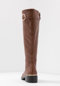 Botte haute marron en similicuir, bout arrondi, talon bloc bas et détail en métal doré sur la bride près du haut.