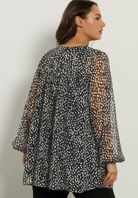 Donna che indossa una blusa nera morbida con macchie bianche irregolari e maniche lunghe trasparenti, vista di spalle contro un muro semplice.