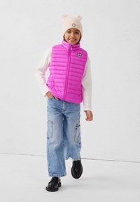 Roze donsjack zonder mouwen met horizontale stiknaden, witte lange mouwen shirt, lichtblauwe cargobroek en zwarte schoenen. Beige beanie met oordetails.