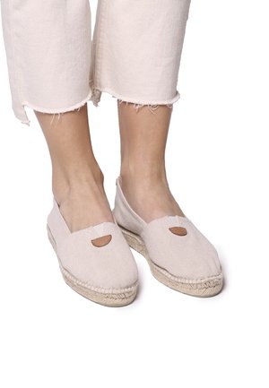 Light beige canvas espadrilles with a flat jute sole, triangular cutouts, and brown leather accents. Worn with cropped, frayed white pants.