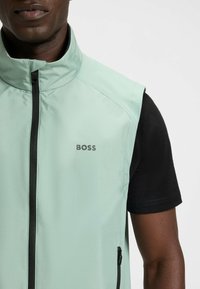 Mintgrünes ärmelloses Jackett mit hohem Kragen, durchgehendem Reißverschluss und schwarzen Akzenten. Verfügt über ein Logo auf der Brust und seitliche Reißverschluss-Taschen.