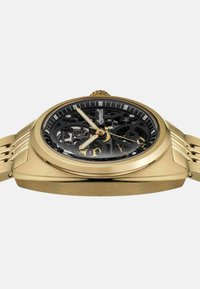 Goudkleurig mechanisch horloge met een zwarte skeletdial, markeringen en gouden wijzers. Gaasband complimenteert het hoekige kastontwerp.