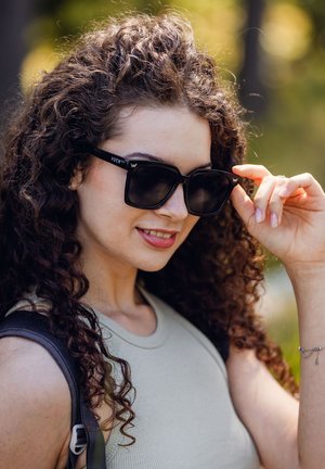 Černé, masivní sluneční brýle s ostrým designem a tmavými čočkami. Na spáncích je logo. Model má kudrnaté vlasy a nosí béžový top.