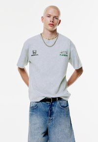 Grå bomulds t-shirt med Honda-logo og NSX-grafik i grønt, parret med løse denimshorts og et sort bælte, lagdelt med halskæder.
