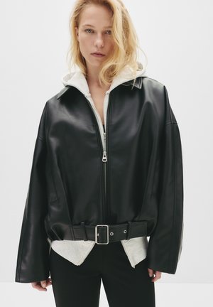 Giacca di pelle nera con colletto alto e zip, dotata di una fodera con cappuccio bianco, vestibilità ampia e una cintura larga in vita.
