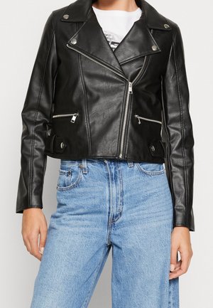 Femme portant une veste courte de motard en cuir noir avec des fermetures éclair argentées et un jean taille haute bleu clair, les mains détendues le long du corps.