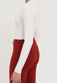 Personne portant un haut blanc côtelé à manches longues, rentré dans un pantalon rouge ajusté taille haute, sur un fond clair uni.