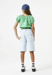 Top corto verde con detalles florales, pantalones cortos azul claro, zapatos en blanco y negro, y una gorra azul. Vista posterior, mangas cortas, dobladillo elástico.
