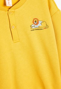 Suéter de algodón amarillo con textura acanalada, que presenta dos botones y una ilustración de un león con una camisa a rayas acompañada del texto "¡BUEN DÍA A RAYAS!"