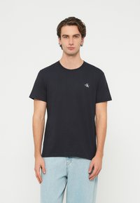 Čierne bavlnené tričko s krátkymi rukávmi, okrúhlym výstrihom a malým bielym logom na ľavej strane hrudníka. Nosené s svetlomodrými džínsami.