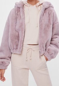 Vrouw in een lichtpaarse fuzzy ritsjack over een crème cropped hoodie en crème joggingbroek met trekkoord, hand in haar zak.