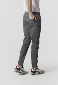 Hombre de pie con la mano en el bolsillo trasero, vistiendo pantalones jogger grises y zapatillas blancas con detalles en negro y rojo.