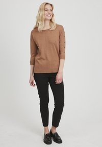 Brązowy dzianinowy top z rękawami o długości trzy czwarte i guzikami, w połączeniu z dopasowanymi czarnymi spodniami i czarnymi mokasynami.