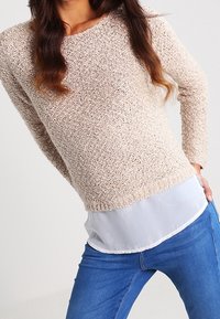 Pull beige en tricot avec une finition texturée, doté d'un col rond, de manches longues, superposé à une chemise blanche légère.