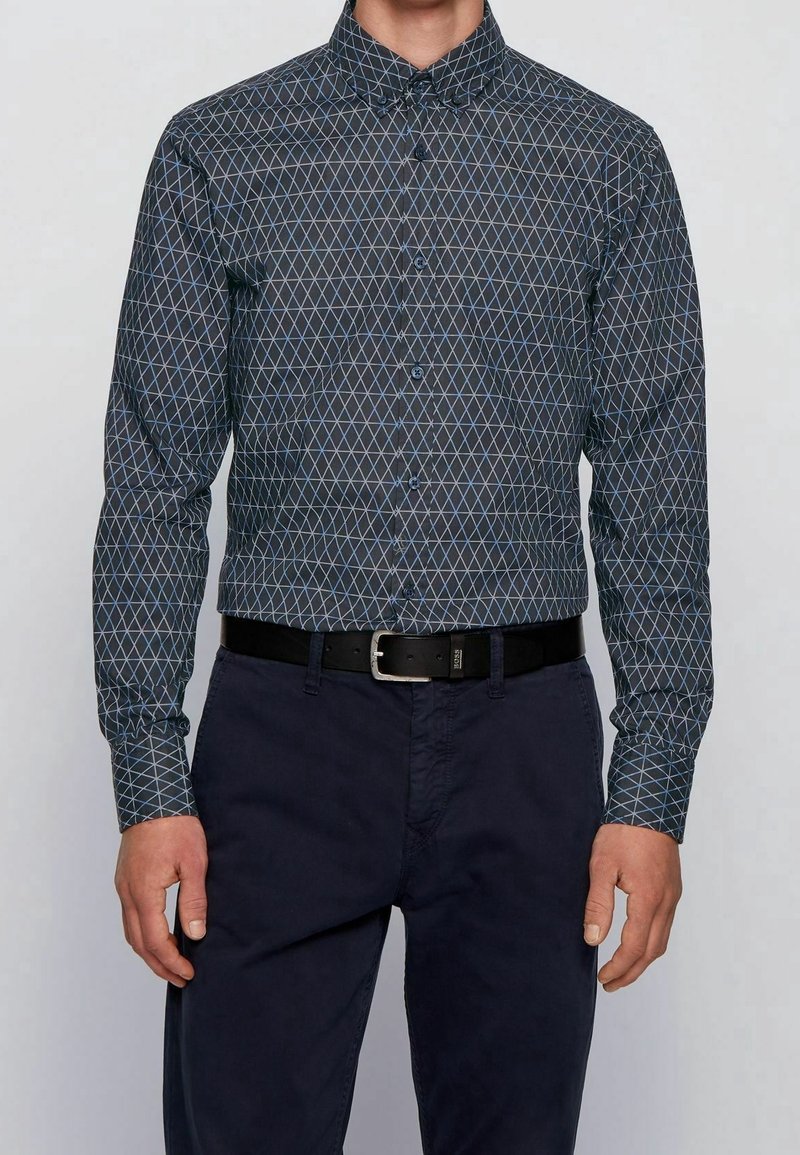 Langarm overhemd met knopen in donkere stof met een diamantpatroon in het blauw. Gecombineerd met donkere broek en een zwarte riem.