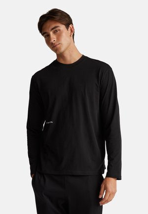 Giovane uomo con capelli corti marroni, vestito con una maglietta nera a maniche lunghe e pantaloni neri abbinati, in piedi davanti a uno sfondo bianco.