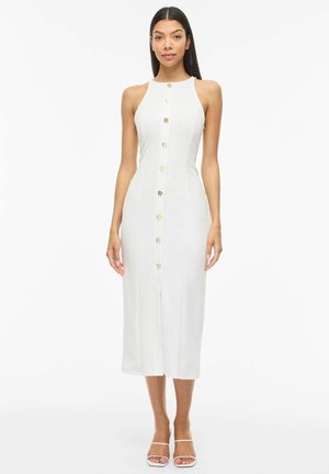 Žena v bielej midi šatách bez rukávov so zlatými gombíkmi vpredu, stojaca s uvoľnenými rukami pozdĺž tela na bielom pozadí.