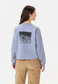 Hellblaues Sweatshirt mit cropped Design, auf der Rückseite mit einem Schwarz-Weiß-Bergmotiv und gerippten Bündchen.