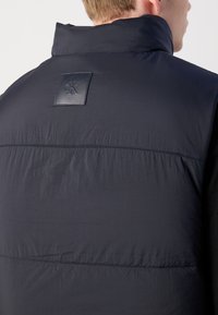 Uomo che indossa un gilet imbottito di colore blu scuro con colletto alto e una patch in pelle nera con il logo "cK" sulla parte superiore della schiena.