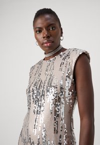 Abito senza maniche coperto di paillettes nei toni beige e argento, con motivi verticali e scollatura arrotondata. Accento con collana choker in cristallo grosso.