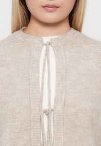 Hellbeige gestrickte Strickjacke mit Rundhalsausschnitt, die eine vordere Bindeschließe und gerippte Akzente aufweist. Weicher Stoff und minimalistische Designmerkmale.