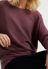 Nainen, jolla on hartiapituinen vaaleanblondi hius, pukeutunut ribbikuvioiseen, pitkähiharaiseen violettiin neuleeseen ja tummiin housuihin, istuu yksi käsi jalan päällä.