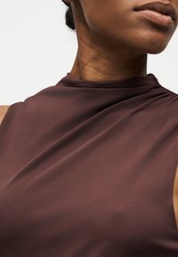 Κοντινό πλάνο ενός ατόμου που φοράει ένα αμάνικο, σκούρο καφέ τοπ με ψηλό λαιμό και λεπτή λεπτομέρεια από draped ύφασμα.