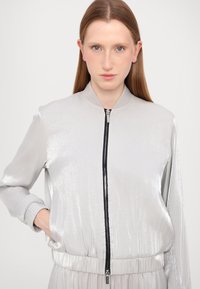 Srebrna bomber jakna sa zatvaračem, rebrastim manžetama i rubom. Refleksivna tekstura, blago skupljena u struku.