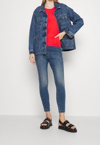 Denimjacka med fickor, röd t-shirt, blå skinny jeans och svarta sandaler med nitar. Avslappnad outfit med lager av texturer och kontrasterande färger.