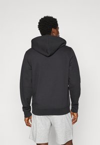 Schwarze Kapuzenjacke mit Kordelzug, gerippten Bündchen und Saum, aus weichem Stoff, über grauen Shorts getragen, von hinten gesehen.