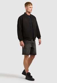Homme portant une veste bomber noire boutonnée, un t-shirt noir, un short en jean noir délavé et des baskets noires, debout devant un fond uni.