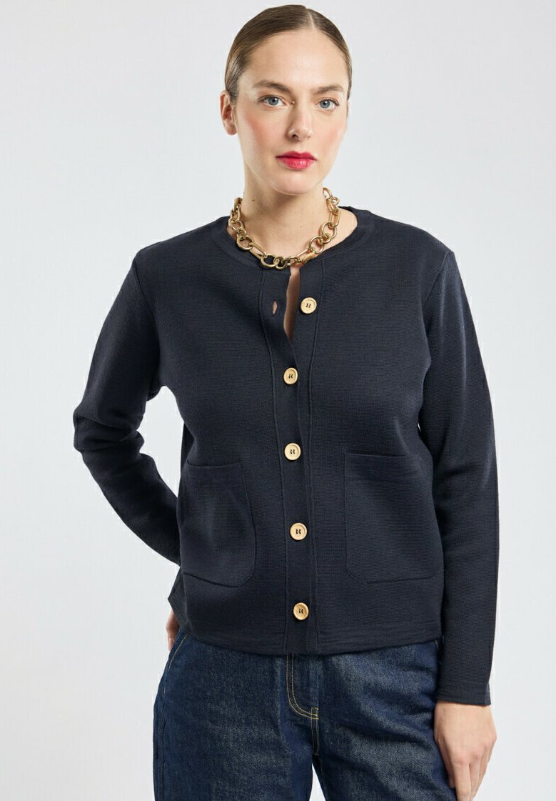 Femme portant un cardigan bleu marine à boutons avec des poches avant, associé à un jean bleu foncé et un collier en chaîne dorée épaisse.