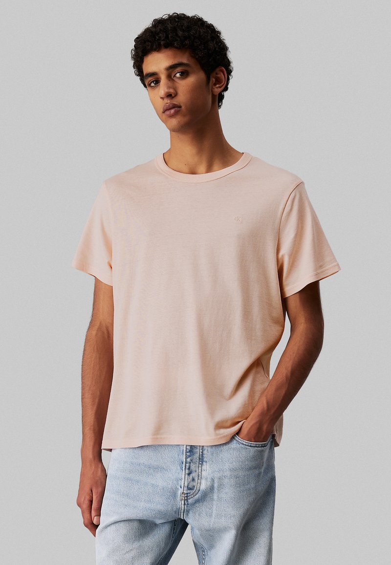 T-shirt en coton pêche clair avec un col rond, des manches courtes et un logo discret sur la poitrine, porté avec un jean en denim bleu clair.