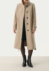 Cappotto in misto lana beige con colletto alto, sei bottoni frontali e maniche con risvolto. Indossato sopra un vestito verde scuro e stivaletti neri.